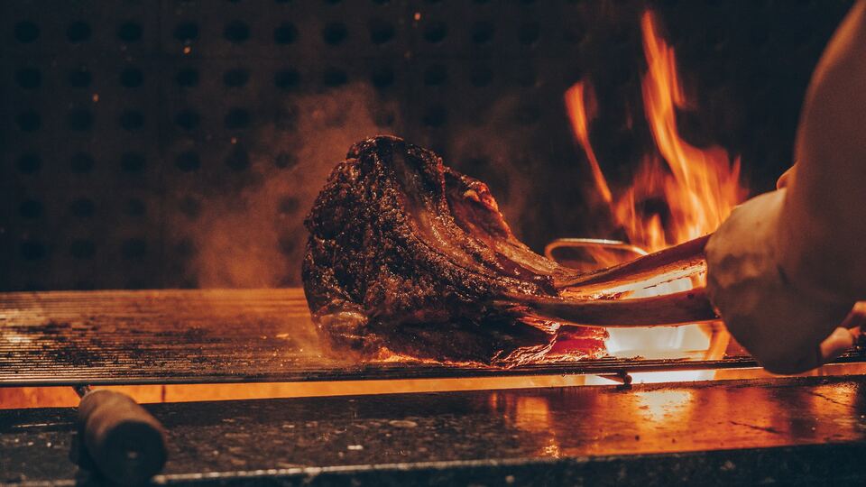 Peça de carne a ser virada na grelha, com chamas ao fundo